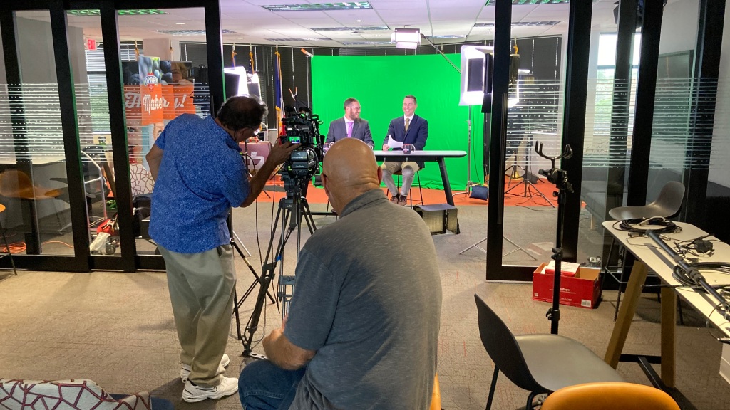 Two person shooting crew recording talent on portable green screen at an office location.